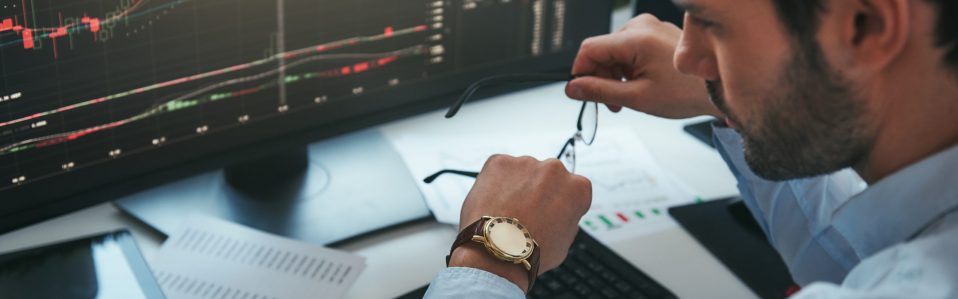 Time to trade. Bearded male trader looking at watch on his hand while working with data and charts on computer screens in his modern office. Stock exchange. Trade concept. Investment concept